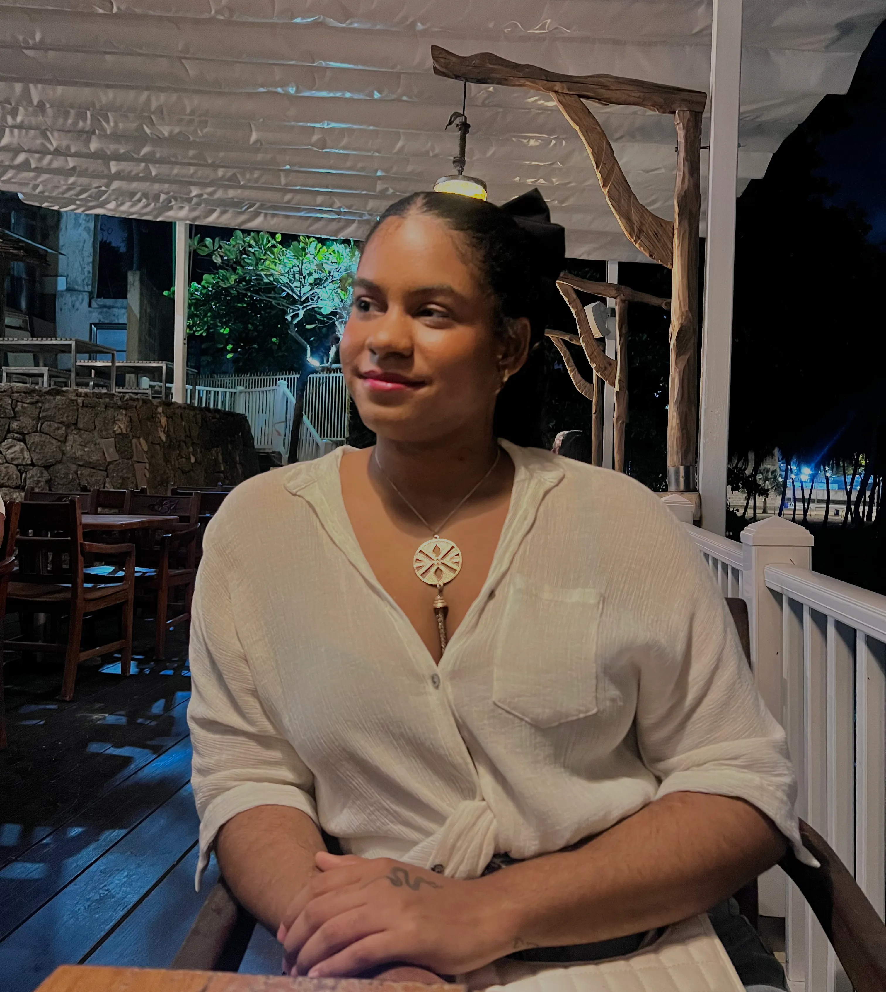 A photo of Maitee Batista smiling gently, dressed in a white tie-front shirt and a circular necklace, seated at a wooden table on a softly lit terrace in the evening.